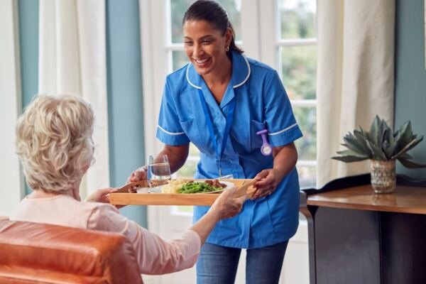 trabalhadora de cuidados feminina em uniforme trazendo refeicao em bandeja para uma mulher idosa sentada na sala de estar em casa 1218270 5172 - Booking-Senior.com